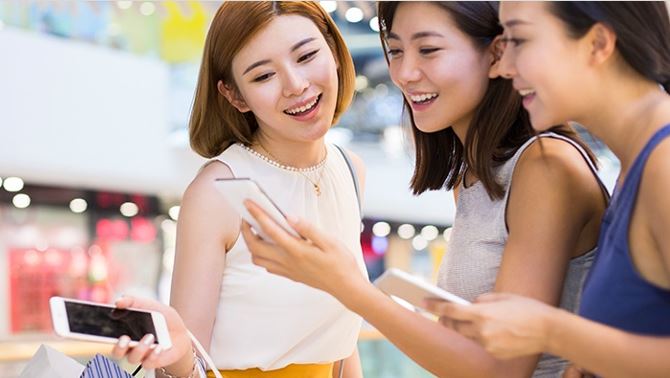Three young women happily checking their mobile phones in a shopping mall; image used for HSBC Macau Pulse UnionPay Dual Currency Diamond Credit Card page